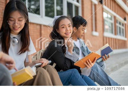 A positive, cute young Asian female college student sits on a footpath with her friends. A positive, cute young Asian female college student sits on a footpath with her friends. 109717086