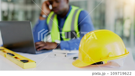 Engineering, construction tools and architect with helmet on desk with break from project, worksite or renovation. African, person or foreman with laptop to review of plans, building or real estate 109719622