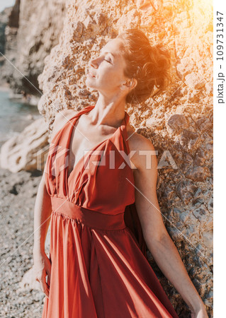 Woman red dress sea. Woman in a long red dress posing on a beach with rocks on sunny day. Girl on the nature on blue sky background. 109731347