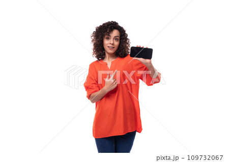 young stylish curly caucasian brunette woman dressed in orange blouse shows phone screen 109732067