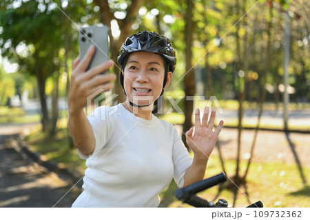 Cheerful middle age woman having video call on mobile phone while resting from cycling in a green park. 109732362