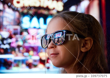Close-up image of little girl in sunglasses standing on street and looking at shop window. Lights reflection on glasses. Big sales 109737075
