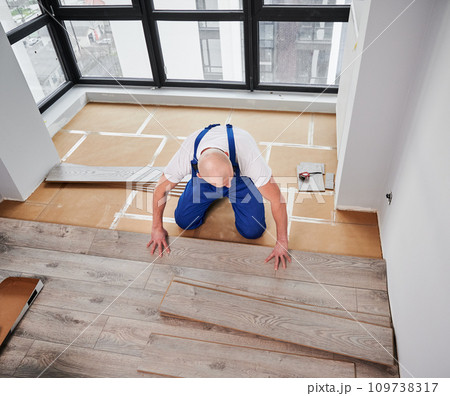 Man laying laminate wood plank on floor underlayment in new flat. Top view of male construction worker installing laminate wooden flooring in living room. Home renovation concept. Man laying laminate wood plank on floor underlayment in new flat. Top view of male construction worker installing laminate wooden flooring in living room. Home renovation concept. 109738317