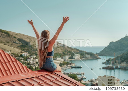 Woman sits on rooftop with outstretched arms, enjoys town view and sea mountains. Peaceful rooftop relaxation. Below her, there is a town with several boats visible in the water 109738680
