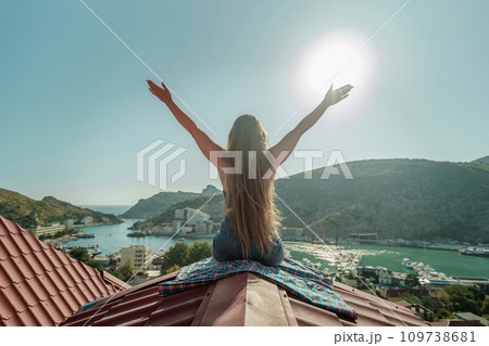 Woman sits on rooftop with outstretched arms, enjoys town view and sea mountains. Peaceful rooftop relaxation. Below her, there is a town with several boats visible in the water Woman sits on rooftop with outstretched arms, enjoys town view and sea mountains. Peaceful rooftop relaxation. Below her, there is a town with several boats visible in the water 109738681