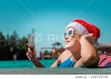 Woman pool Santa hat. A happy woman in a blue bikini, a red and white Santa hat and sunglasses poses near the pool with a glass of champagne standing nearby. Christmas holidays concept. Woman pool Santa hat. A happy woman in a blue bikini, a red and white Santa hat and sunglasses poses near the pool with a glass of champagne standing nearby. Christmas holidays concept. 109738706