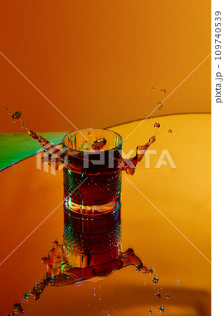 Ice cube falling into glass with strong drink. whiskey and coke against orange background in neon light 109740539
