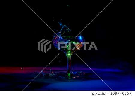 Martini splash. Glass with popular cocktail standing against dark background with neon light with smoke Martini splash. Glass with popular cocktail standing against dark background with neon light with smoke 109740557
