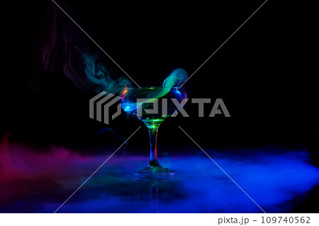 Alcohol splash. Glass with delicious alcoholic drink, popular cocktail with martini against dark background with neon light with smoke 109740562
