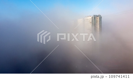A white-and-blue multi-storey residential building is shrouded in fog in the early spring morning. 109741411