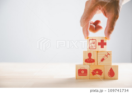 Conceptual portrayal, Hand clutches wooden block with healthcare and medical icons. Reflecting safety, health, and family well-being. Symbolic of pharmacy, heart care, and happiness. Conceptual portrayal, Hand clutches wooden block with healthcare and medical icons. Reflecting safety, health, and family well-being. Symbolic of pharmacy, heart care, and happiness. 109741734