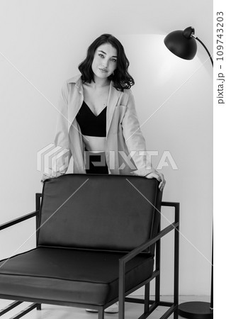 Beautiful young girl in black trousers and a blue jacket near a chair on a white background. Portrait of a beautiful young woman 109743203