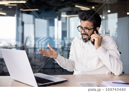 Angry young Indian male businessman sitting in the office at the desk and emotionally talking on the phone. Angry young Indian male businessman sitting in the office at the desk and emotionally talking on the phone. 109743480