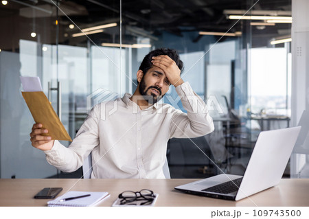 Portrait of a young Indian man who works in the office with a laptop, holds his head with his hand, looks at the camera in shock, received bad news by letter. 109743500
