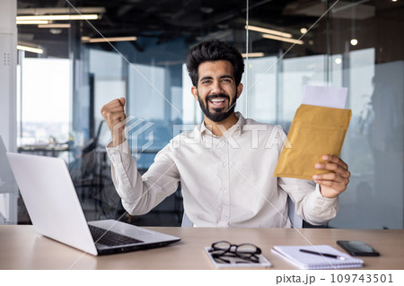 Portrait of a happy Muslim businessman sitting in the office at the table, holding an envelope with a letter in his hands, and rejoices at the camera with success and received news. 109743501