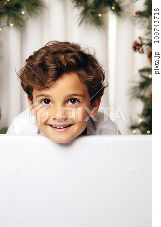 AI-generated content. Cute boy peeking out from behind a white empty board. Christmas promotional banner mockup with copy space and blurred Christmas lights on the background. A child in a cozy 109743718