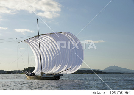 快晴の下、穏やかな霞ヶ浦の湖面に浮かぶ、白い帆をいっぱいに張った帆引き船と遠くに見える筑波山 109750066