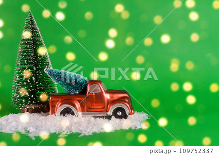 A red pickup truck is carrying a Christmas tree with artificial snow on a green background with bokeh. Banner, New Year card with copy space 109752337