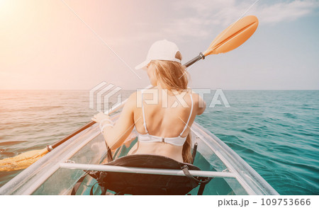Woman in kayak back view. Happy young woman with long hair floating in transparent kayak on the crystal clear sea. Summer holiday vacation and cheerful female people relaxing having fun on the boat Woman in kayak back view. Happy young woman with long hair floating in transparent kayak on the crystal clear sea. Summer holiday vacation and cheerful female people relaxing having fun on the boat 109753666