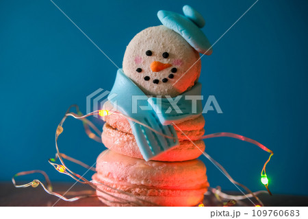 Snowman macaron dessert decorated with hat and scarf wrapped with colorful decorative garland lights against blue background. Merry Christmas and Happy New Year festive concept 109760683