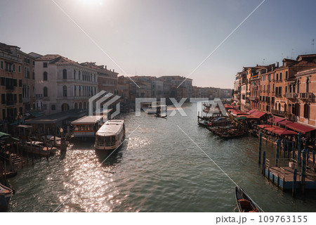 Experience the vibrant and lively atmosphere of the grand canal in Venice, Italy. This sunny scene captures locals, tourists, and gondolas against a neutral color palette. 109763155