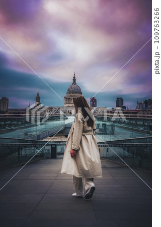 Female tourist walking on bridge while looking at St. Paul's cathedral. Woman wearing coat exploring religious building with cloudy sky in background. She is enjoying vacation in London. 109763266