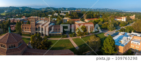 Aerial view of UCLA campus with Gothic tower, red-brick buildings, green lawns, and pathways amidst hilly, tree-covered landscape in soft, golden light. Aerial view of UCLA campus with Gothic tower, red-brick buildings, green lawns, and pathways amidst hilly, tree-covered landscape in soft, golden light. 109763298
