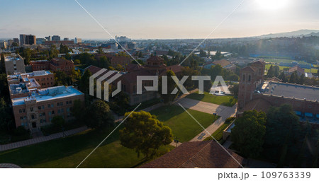 Aerial view of a university campus at sunrise or sunset, featuring historic red-brick buildings, modern architecture, lush greenery, and a city backdrop under a clear sky. 109763339