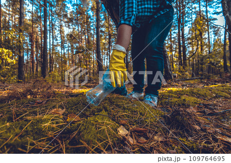 A hand in a yellow glove picks up a plastic bottle from green moss in the forest and puts it in a trash bag. A woman eco-defender collects garbage in the forest, saving it from pollution. 109764695