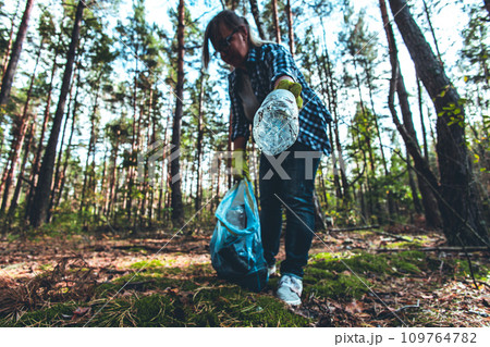 A woman with a garbage bag collects plastic garbage in the autumn forest. Saving the planet from garbage. A woman wearing protective gloves fights to save the planet from garbage. A woman with a garbage bag collects plastic garbage in the autumn forest. Saving the planet from garbage. A woman wearing protective gloves fights to save the planet from garbage. 109764782