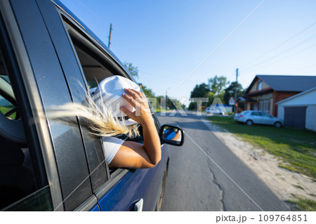 Beautiful woman with flying hair in the car window while driving. Feeling of freedom. In summer, while traveling by car, a blonde woman leaned out the window and enjoys freedom and vacation. 109764851