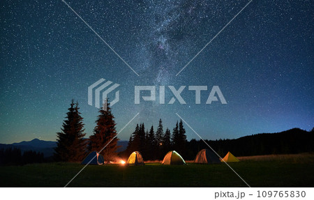 Night camping in mountains under starry sky. Tourist tents in campsite near burning campfire under beautiful sky full of stars with Milky way above forest. Concept of tourism and traveling. Night camping in mountains under starry sky. Tourist tents in campsite near burning campfire under beautiful sky full of stars with Milky way above forest. Concept of tourism and traveling. 109765830