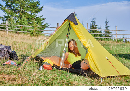 Woman traveller camping outdoors. Female tourist traveling, hiking in mountains. Attractive, sporty woman sitting in tent, having break, looking at camera. Concept of tourism and adventure. 109765839