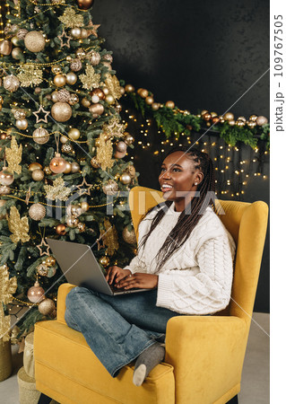 Beautiful young african female sitting with laptop near Christmas tree at home 109767505