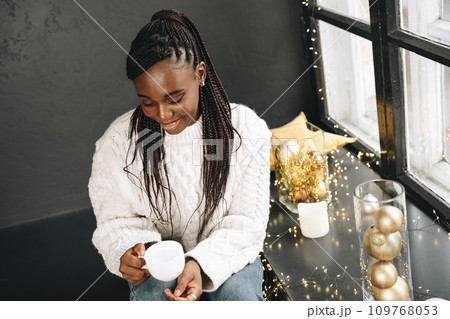 African woman in warm white sweater drinking hot coffee or tea at home African woman in warm white sweater drinking hot coffee or tea at home 109768053
