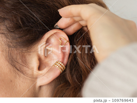 Cropped close-up shot of a young woman with two asymmetrical golden ear cuffs. Female with golden ear cuffs, side view. Cropped close-up shot of a young woman with two asymmetrical golden ear cuffs. Female with golden ear cuffs, side view. 109769643