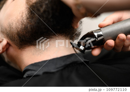 A barber in the salon trims the beard of a man with a trimmer in the neck area 109772110