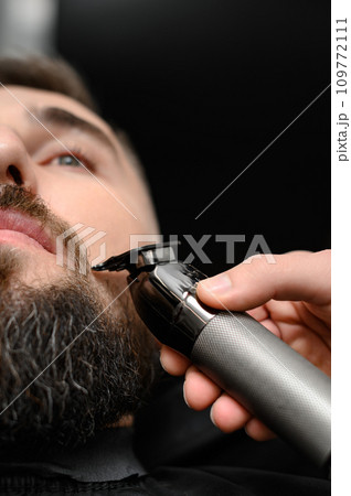 Cutting a gentlemans beard in a barbershop with a clipper. Shortening the length of the beard from the sides by the master for the client. Cutting a gentlemans beard in a barbershop with a clipper. Shortening the length of the beard from the sides by the master for the client. 109772111