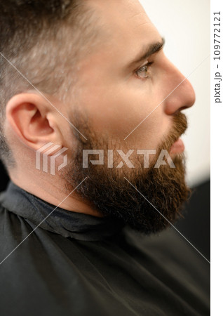 A young man with a beard in a black cape sits on a chair in a barbershop. The client is waiting for a haircut of the beard. A young man with a beard in a black cape sits on a chair in a barbershop. The client is waiting for a haircut of the beard. 109772121