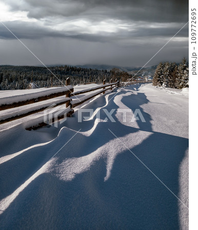 Picturesque waved shadows on snow from wood fence. Alpine mountain winter hamlet outskirts, snowy path, fir forest. High resolution image with great depth of field. 109772695