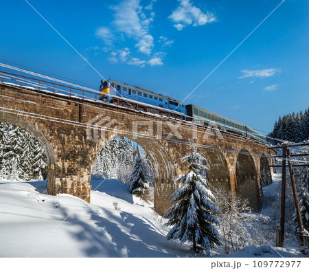 Stone viaduct (arch bridge) on railway through mountain snowy fir forest and locomotive  with a passenger train. Snow drifts  on wayside and hoarfrost on trees and electric line wires. 109772977