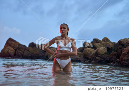 Attractive woman enjoys nighttime swim in ocean under crimson illumination. Fashionable lady in swimwear poses during an evening ocean dip. Attractive woman enjoys nighttime swim in ocean under crimson illumination. Fashionable lady in swimwear poses during an evening ocean dip. 109775254