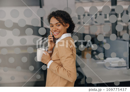 Pretty female freelancer with coffee cup is talking phone while standing on coworking background 109775437