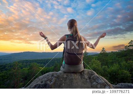 Sportive female hiker sitting alone taking a meditation break on hillside trail. Lonely woman enjoying view of evening nature from rocky cliff on wilderness path. Active lifestyle concept Sportive female hiker sitting alone taking a meditation break on hillside trail. Lonely woman enjoying view of evening nature from rocky cliff on wilderness path. Active lifestyle concept 109776271