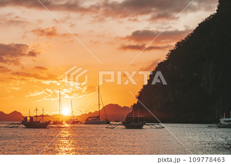 Traditional philippine boats bangka at sunset time. Beautiful sunset with silhouettes of philippine boats in El Nido, Palawan island, Philippines. Orange sky sunset paradise beach. Traditional philippine boats bangka at sunset time. Beautiful sunset with silhouettes of philippine boats in El Nido, Palawan island, Philippines. Orange sky sunset paradise beach. 109778463