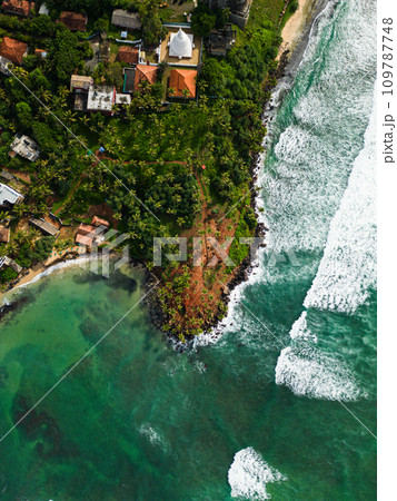 Aerial view from above of lush tropical coastal landscape and vibrant turquoise sea with white-capped waves. Tranquil bay, a traditional village temple, exotic travel destinations nature beauty. 109787748