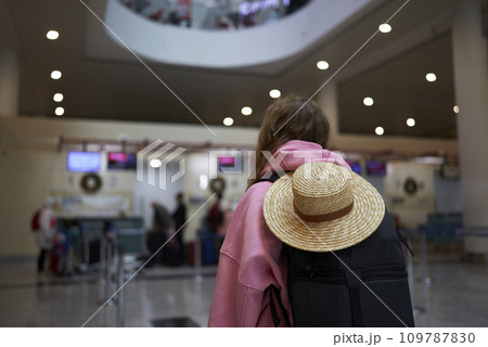 Back view of a traveler with a sun hat and backpack at the airport during holiday season, festive vacation departure. Back view of a traveler with a sun hat and backpack at the airport during holiday season, festive vacation departure. 109787830
