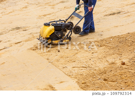Worker using vibratory plate compactor for compaction sand during path construction. 109792314
