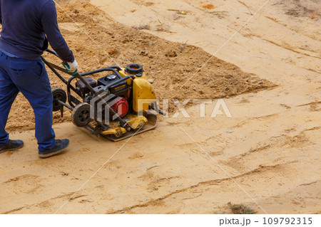 Worker using vibratory plate compactor for compaction sand during path construction. 109792315