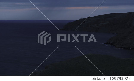 Aerial view of the long grey cliff covered with shrubs and the lighthouse near sea against blue with pink evening sky. Shot. Beautiful evening marine scenery Aerial view of the long grey cliff covered with shrubs and the lighthouse near sea against blue with pink evening sky. Shot. Beautiful evening marine scenery 109793922